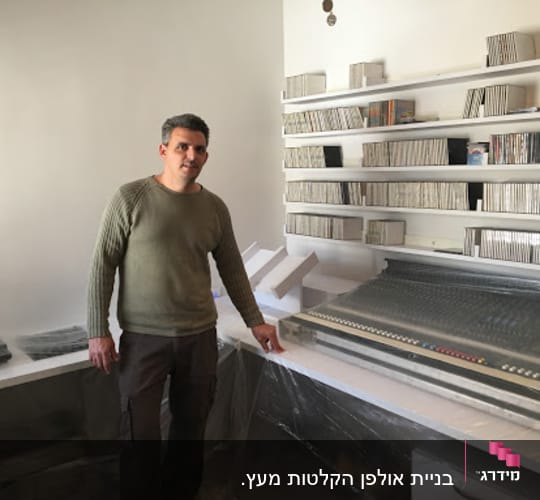 A man standing next to a large audio mixing console in a room with shelves filled with CDs and audio equipment.